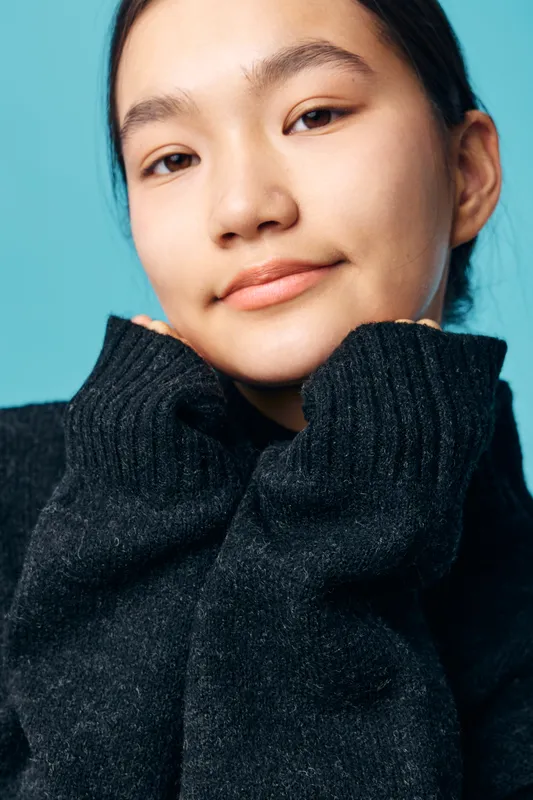 Young woman wearing black sweater with hands near face, looking at camera with calm expression on blue background. Casual portrait of female model with natural makeup, relaxed pose and soft lighting.