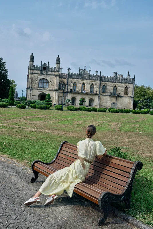 A woman in a vintage dress sits on a wooden bench in a manicured park, facing a grand historic castle, creating a serene, timeless scene.