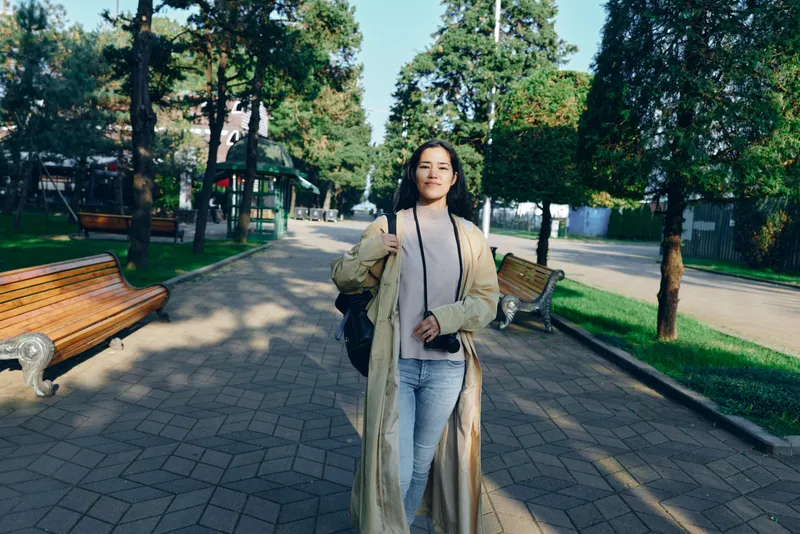 Woman in a beige coat walking along a park pathway beneath leafy trees, casual street style, sunlight, and a peaceful urban park backdrop during a daytime outdoor shoot