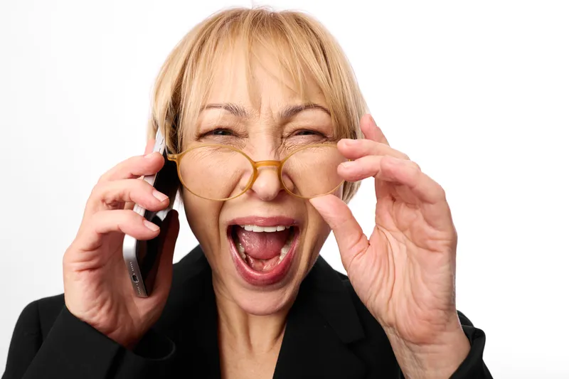 Surprised mature woman in studio portrait, holding a smartphone to her ear and adjusting glasses, open mouth shout, white background, business communication