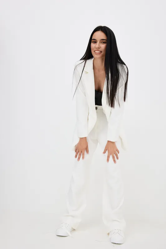 Young woman smiling, dressed in white pantsuit and black top, standing with hands on knees against minimalist white background, long black hair flowing.