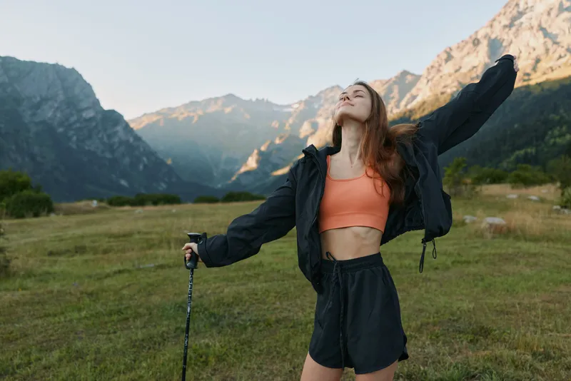 A joyful outdoors enthusiast stands in a meadow surrounded by mountains, arms raised, wearing a coral crop top and dark windbreaker, savoring fresh air and freedom under clear skies.