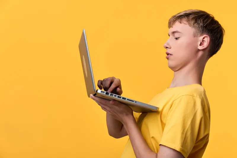 Boy in a yellow shirt using a laptop against a bright yellow background, focused on the screen, illustrating technology, learning, and online work daily.