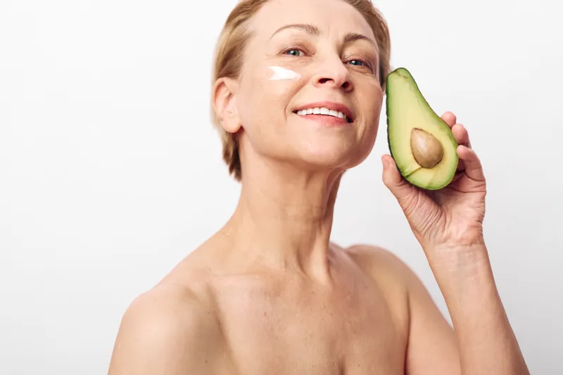 Smiling mature woman with natural skin care cream on face holds fresh avocado, showing healthy beauty and wellness concept on white background with soft lighting for skincare routine.