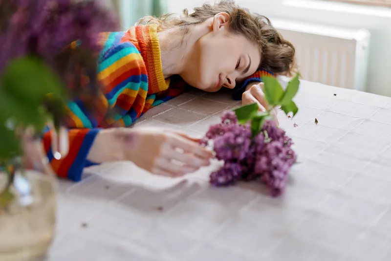 Young woman with a striped colorful sweater rests her head on a table while writing beside blooming flowers. A cozy home lifestyle moment focused on creativity and calm.