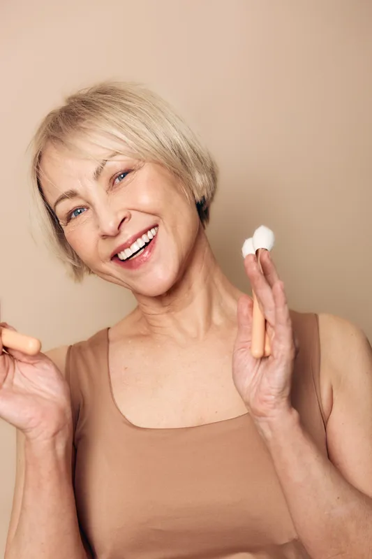 Happy middle aged woman with short blonde hair holds cotton pad and makeup brush smiling in beige top on neutral background. Concept of skincare, beauty routine and natural care.