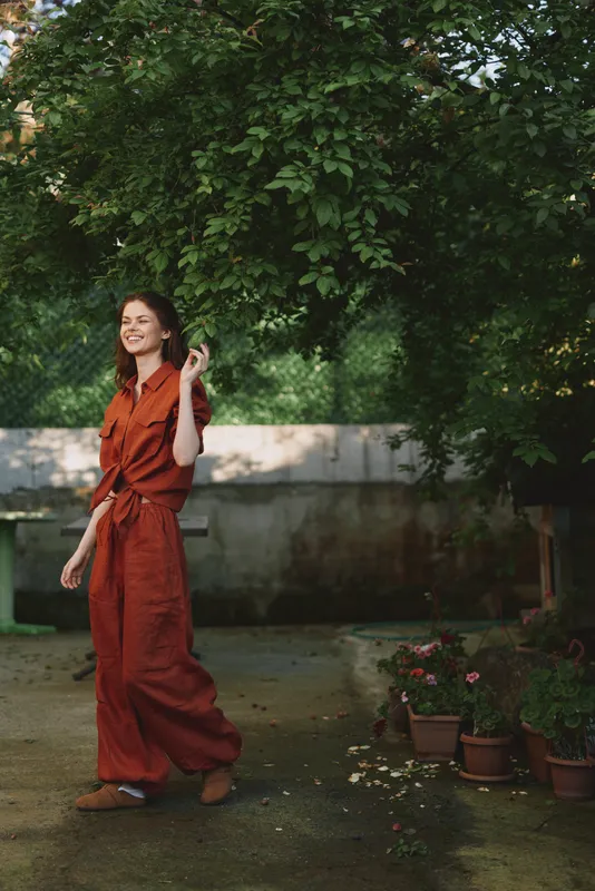 Woman in rust jumpsuit enjoys outdoor garden setting, expressing joy with a bright smile, candid lifestyle moment, casual fashion, natural light, peaceful surroundings