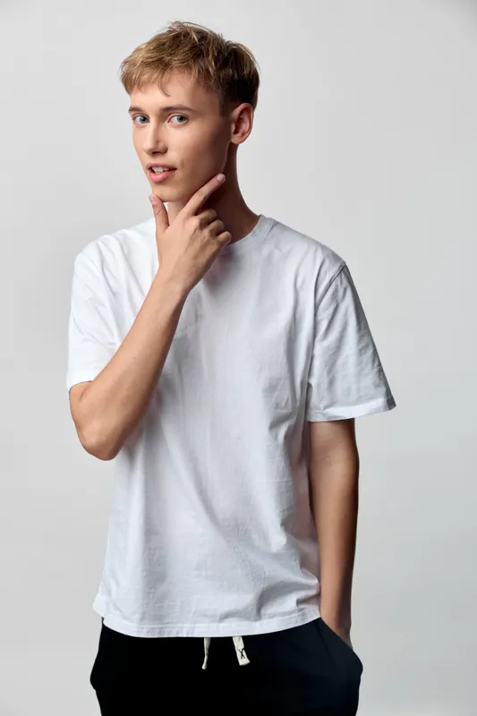 Thinking man in casual white t shirt stands in studio with thoughtful expression, calm mood and relaxed pose, highlighting minimalism lifestyle and fashion on a gray background