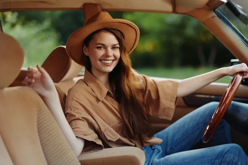 Happy young woman driving a vintage car with a stylish hat, showcasing a casual fashion style in a natural outdoor setting, embodying freedom and joy.