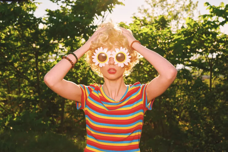Playful young woman outdoors wears sunflower goggles and a striped shirt, posing with hands on her head in bright sunlight for a carefree, energetic vibe.
