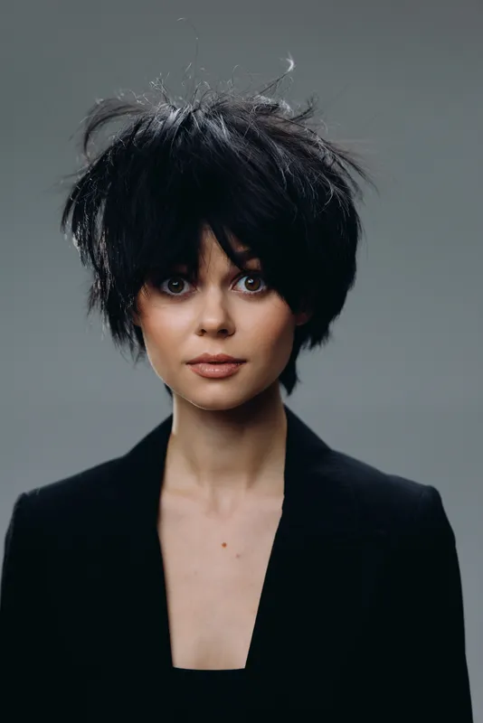 Portrait of a young woman with messy black hair wearing a black jacket and neutral expression against a gray background.