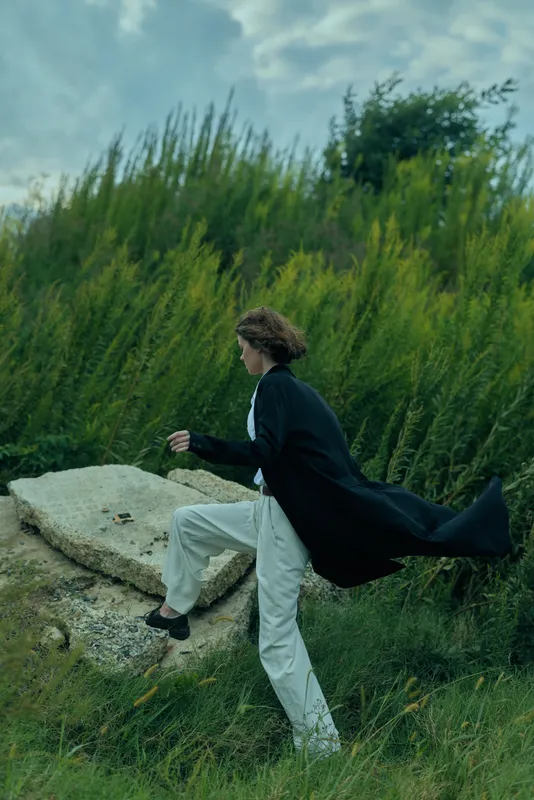 Individual in a dark cape and light trousers strides through a grassy landscape, capturing motion and fashion in a dynamic outdoor scene with a natural backdrop and wind swept energy