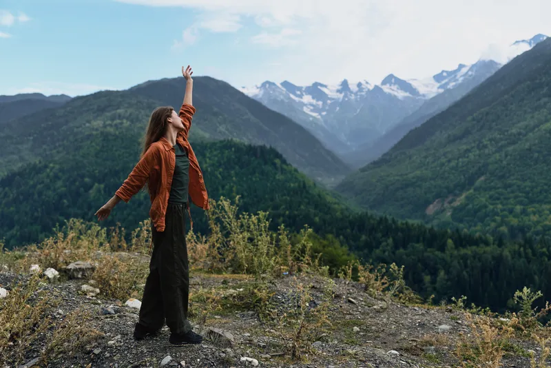 Hiking woman stands on a rocky overlook with arms raised, enjoying nature and a sweeping mountain landscape, expressing adventure, freedom, and joy in the outdoor setting