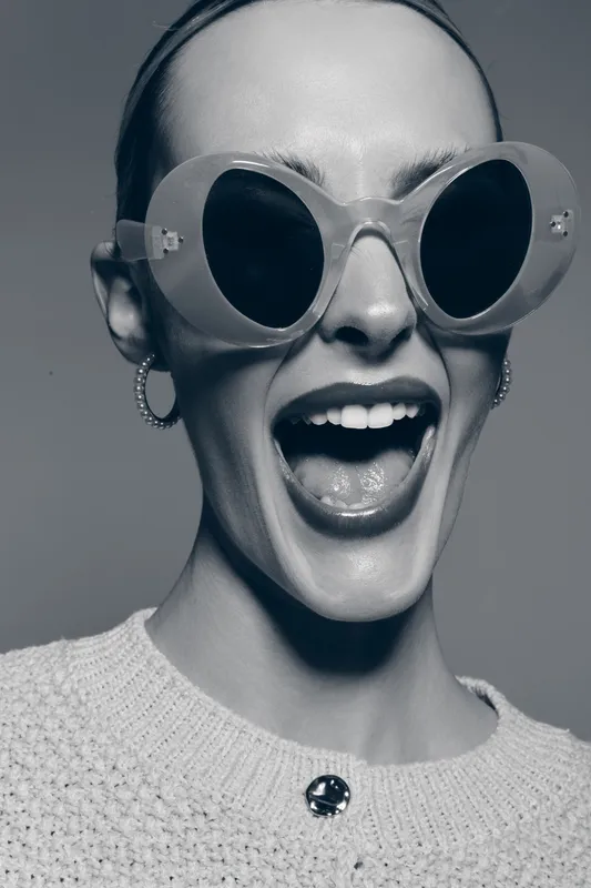Happy Caucasian woman with light skin wearing oversized round sunglasses and hoop earrings smiles widely. Monochrome studio portrait with textured sweater and joyful expression.