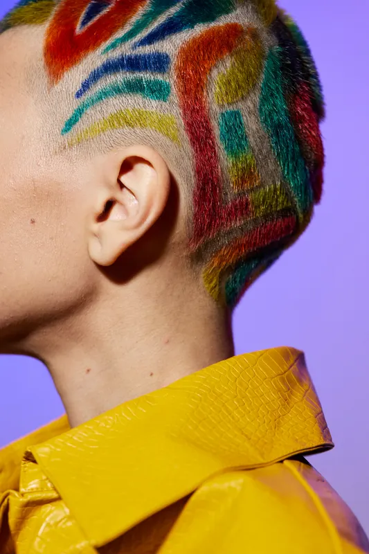Woman with colorful painted hair and shaved sides wearing a bright yellow textured jacket against a vibrant purple background, showcasing creative self-expression and bold style.