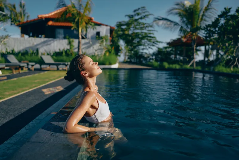 Young woman in a white swimsuit relaxing in an outdoor pool near tropical plants and villas. Lifestyle concept with natural sunlight and calm atmosphere during daytime.