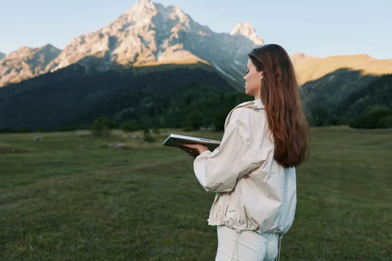 A thoughtful woman stands in a scenic mountain meadow, holding a tablet and looking toward the distant peaks. The calm outdoor setting inspires focus, exploration, and creative work.