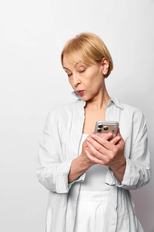 Elderly woman in laboratory coat holds a smartphone, with concerned and worried expression, anxiety and stress shown in a studio portrait on a white background