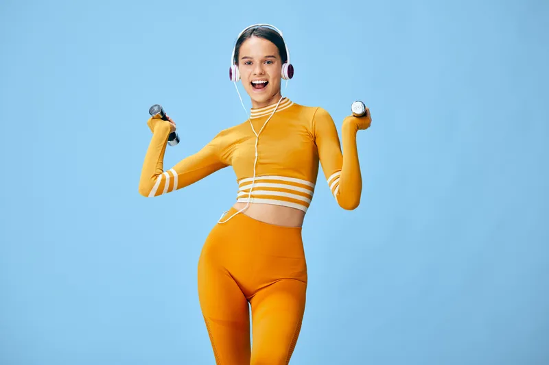 Dynamic woman fitness enthusiast in bright orange outfit lifting dumbbells with headphones, mid workout pose against blue backdrop, conveying energy, motivation, and active lifestyle through bold