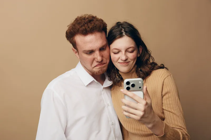 Young couple taking a selfie together with a smartphone. The woman smiles gently while the man makes a funny face. Casual clothing and neutral background create a warm, intimate mood.