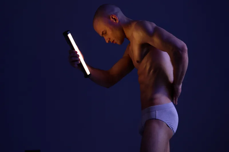 Athletic man exploring lighting techniques in a dimly lit studio, showcasing the interplay of shadows and illumination on his sculpted physique.