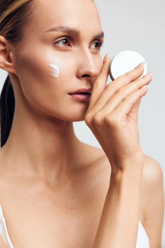 A young woman with clear skin applies face cream from a white jar, focusing on skincare and beauty routine in a bright studio setting with neutral background and soft lighting.