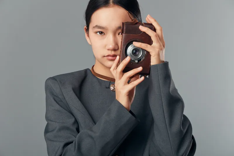 Woman in gray jacket holding vintage camera in front of face posing for photo studio portrait with neutral background and focused expression on creative photography concept.