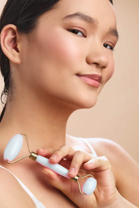 Asian woman uses a facial roller in a serene home setting, enjoying a relaxing skincare moment. The calm expression and soft lighting convey wellness, self care, and a peaceful beauty routine.
