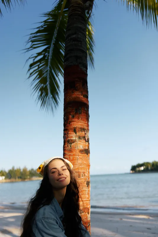 A woman stands beside a tall palm on a sunlit beach, wearing a denim jacket with a relaxed smile, creating a calm tropical portrait against a clear blue sky.