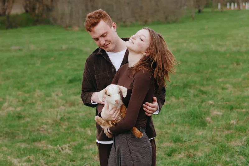 Cheerful couple outdoors, embracing a playful goat kid in a sunlit field, warm breeze, casual clothing, intimate moment, soft colors, natural setting, countryside vibe, candid relationship