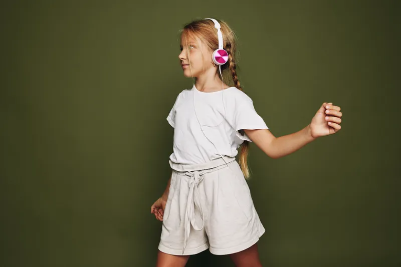 Young girl with braided hair wearing white t-shirt and shorts listens to music with pink headphones. Casual studio shot on olive background.