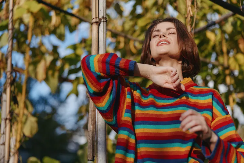 Woman in colorful striped sweater portrait outdoors, basking in warm sunlight with closed eyes, relaxed expression and autumn foliage background creating cozy seasonal mood.