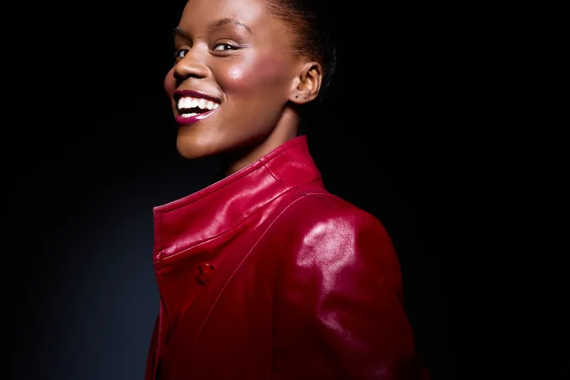 Smiling dark skinned African woman in red leather jacket looks confident and happy against black background. Concept of fashion, beauty, confidence, and positive mood in studio lighting.