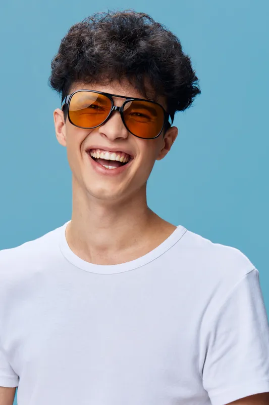 smiling man wearing orange sunglasses and white t-shirt with curly hair on blue background, happy young adult expressing joy and positive energy