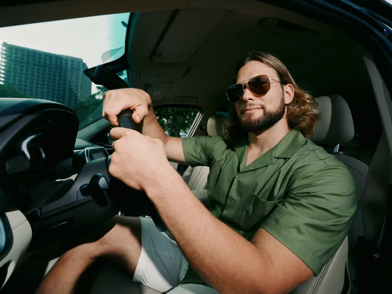 A confident man drives a car, gripping the wheel with both hands, wearing sunglasses and a green shirt, captured inside a modern vehicle cabin under soft daylight.