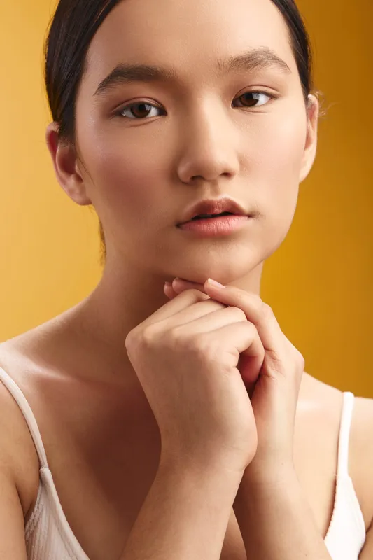 Asian woman gazes softly at the camera with a thoughtful expression, hands gently folded under the chin, warm studio lighting creating a serene, elegant portrait.
