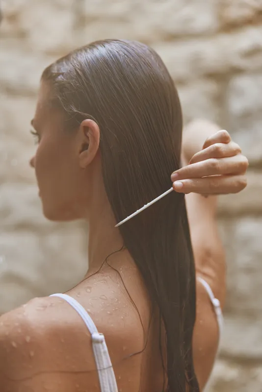Woman combing wet hair with a white comb against soft beige stone wall background, showcasing smooth brown hair and natural skin tones in a serene moment.