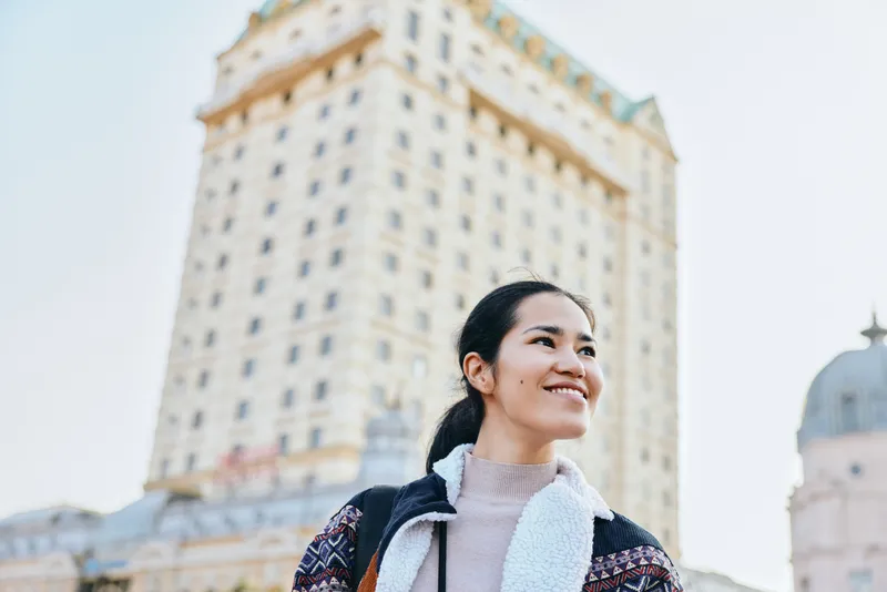 Smiling woman in a chic jacket stands outdoors with a city skyline behind her, capturing a vibrant urban moment, exploration and confidence in a modern street photography style.