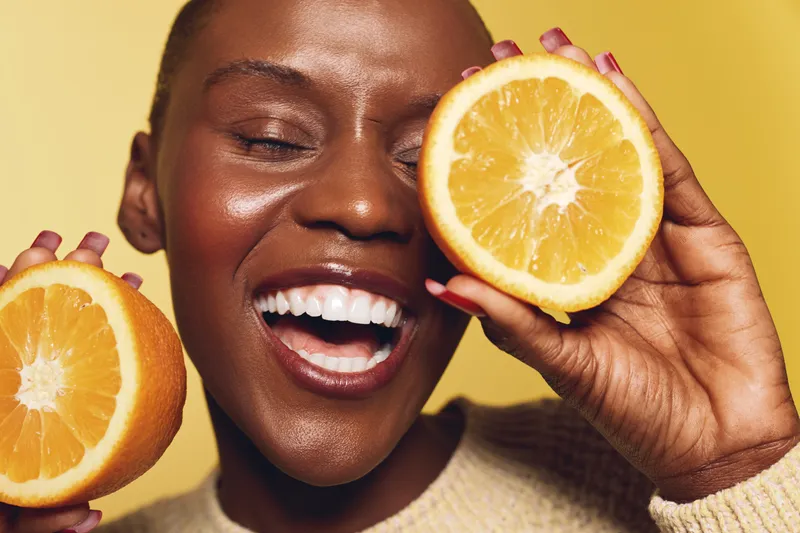Happy African woman with dark skin holds two orange halves near face smiling with closed eyes on yellow background. Concept of fresh fruit, healthy lifestyle, and natural beauty.
