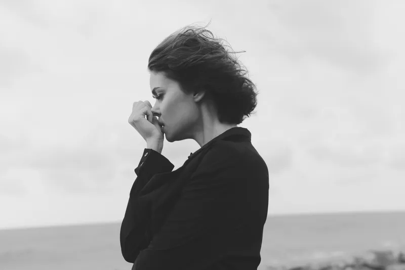 Woman standing by the sea in a dark jacket, hair blown by wind, contemplative pose and moody atmosphere in a minimalist black and white scene with no branding