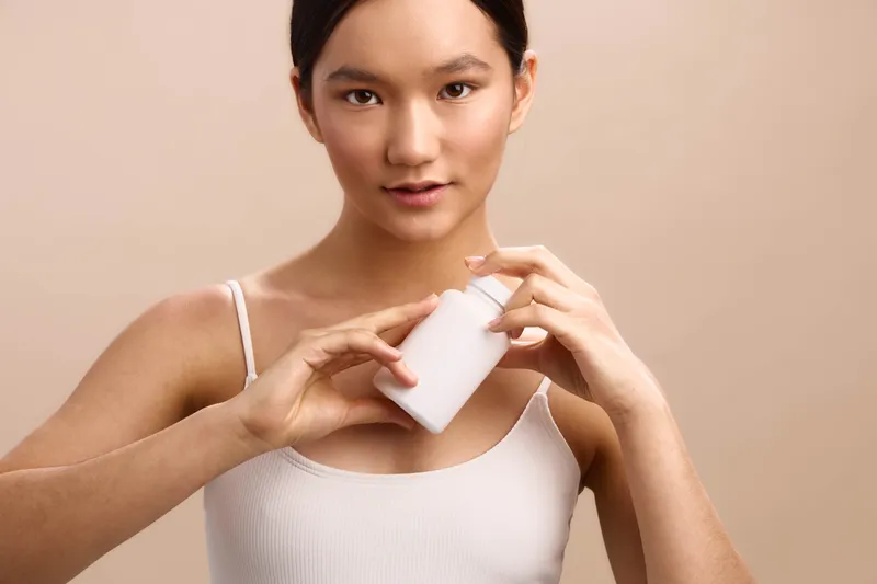 Asian woman in a white tank top holds a small bottle, presenting a skincare routine in a clean studio backdrop with calm lighting and a confident, approachable mood.