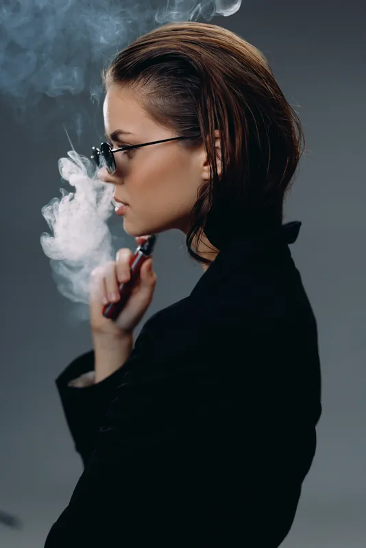 Profile of a thoughtful woman exhaling vapor, wearing glasses and dark clothing, against a dark smoky background.