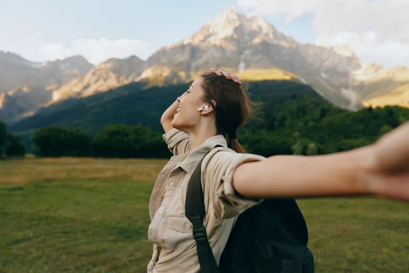 Cheerful woman outdoors, arms outstretched, backpack traveler enjoying sunlit mountains, open field, fresh air, natural colors, relaxed mood, adventurous spirit, scenic landscape