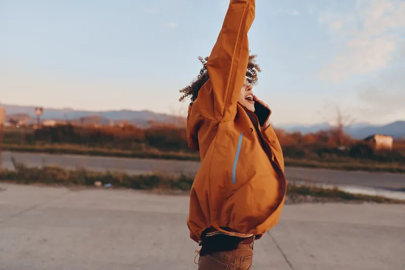 woman lifestyle outdoors, orange jacket with hood up, standing on road at sunset, looking toward mountains, windy evening, soft light, casual travel mood, from behind
