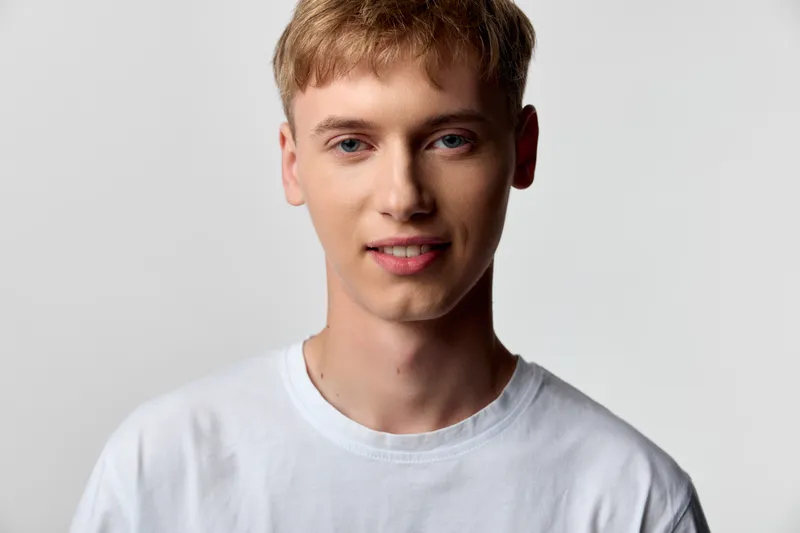 Young man wearing a tshirt stands in a bright studio, smiling, casual and approachable, with clean lighting, neutral background, and a minimalist aesthetic for lifestyle or fashion projects.
