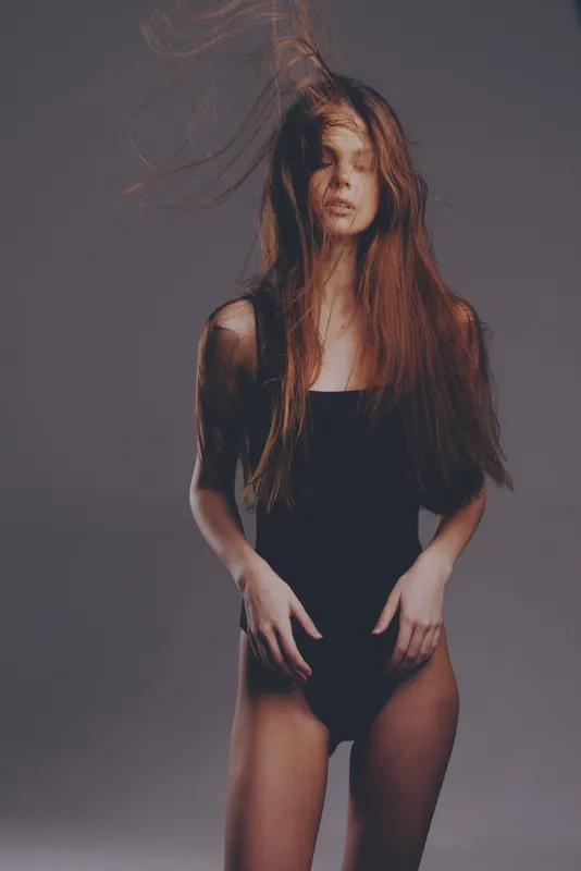 Young woman with long red hair wearing a black swimsuit posing against gray studio background, showing natural beauty and soft expression.
