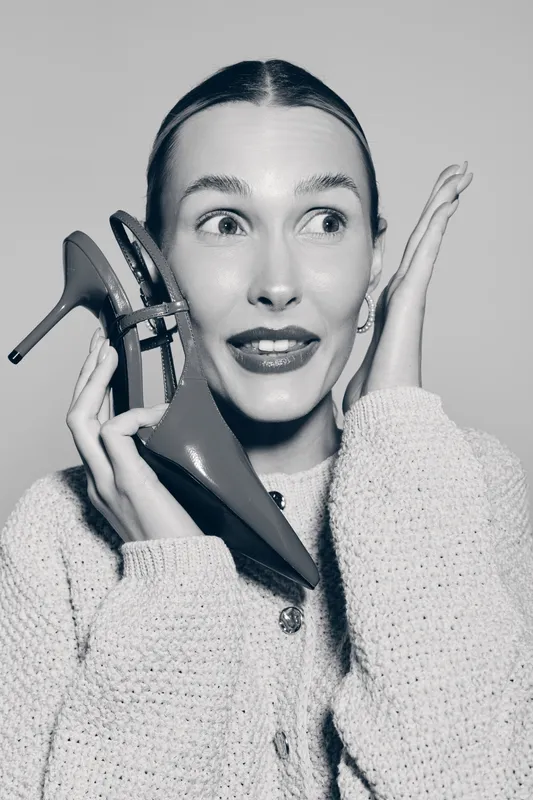 Young white woman with light skin holds red high heel near face with surprised expression. Studio portrait in cozy sweater on light background.