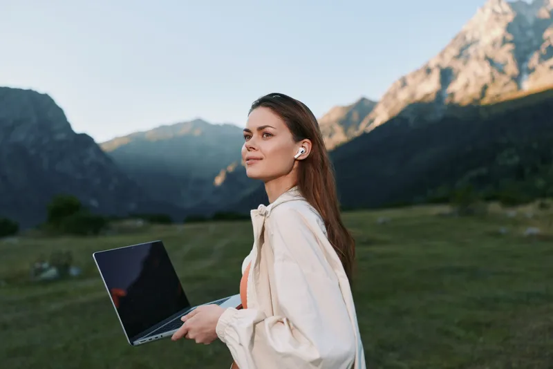 A cheerful professional woman stands outdoors in a mountainous landscape, holding a laptop. She smiles warmly, projecting a calm, productive mood and a modern, nature infused work scene.