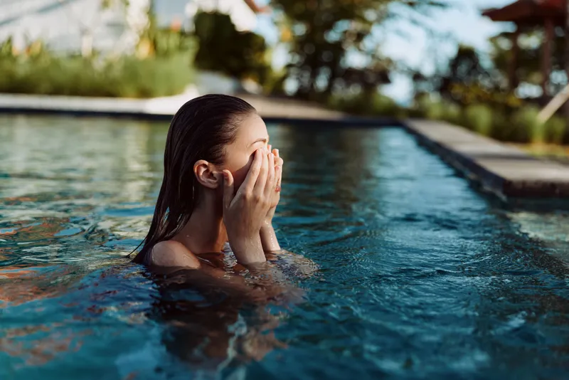 Outdoor pool moment, a woman partially submerged, hands over her face, candid privacy, sunlight glow, clear water, breeze, relaxed posture, vibrant colors, casual summer mood, carefree atmosphere