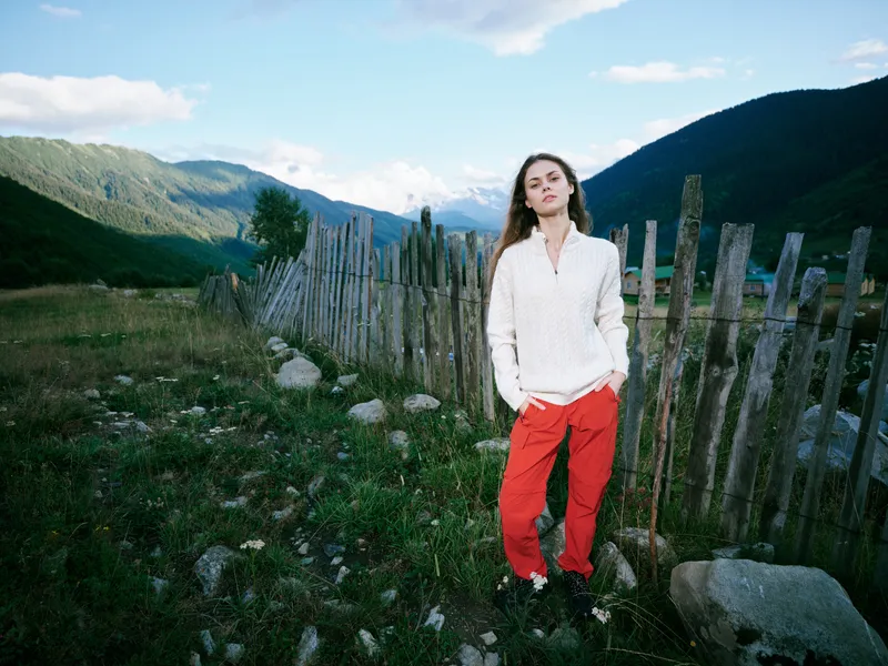 A woman stands in a meadow beside a rustic fence, outdoor scenery with mountains in the distance, blue sky above. Casual clothing and relaxed pose highlight nature, travel and countryside calm